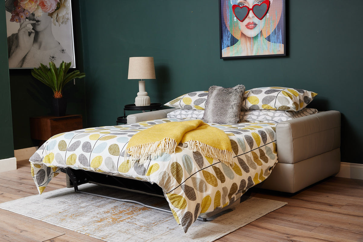 Modern sofa bed extended into a full bed with patterned bedding, a yellow throw, and decorative pillows in a stylish room with artwork and warm wood flooring.