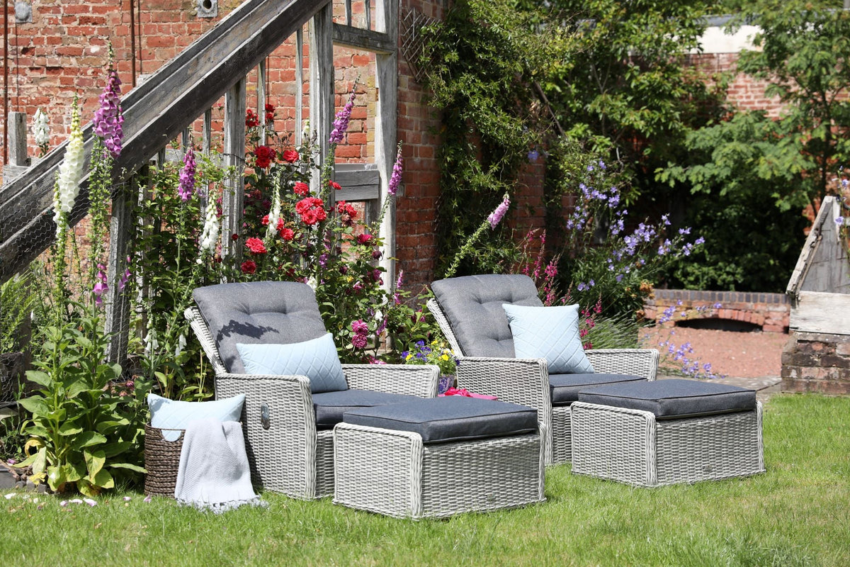 Two grey wicker garden lounge chairs with matching footstools and cushions, arranged on a lawn beside colourful flowers and a rustic brick wall.